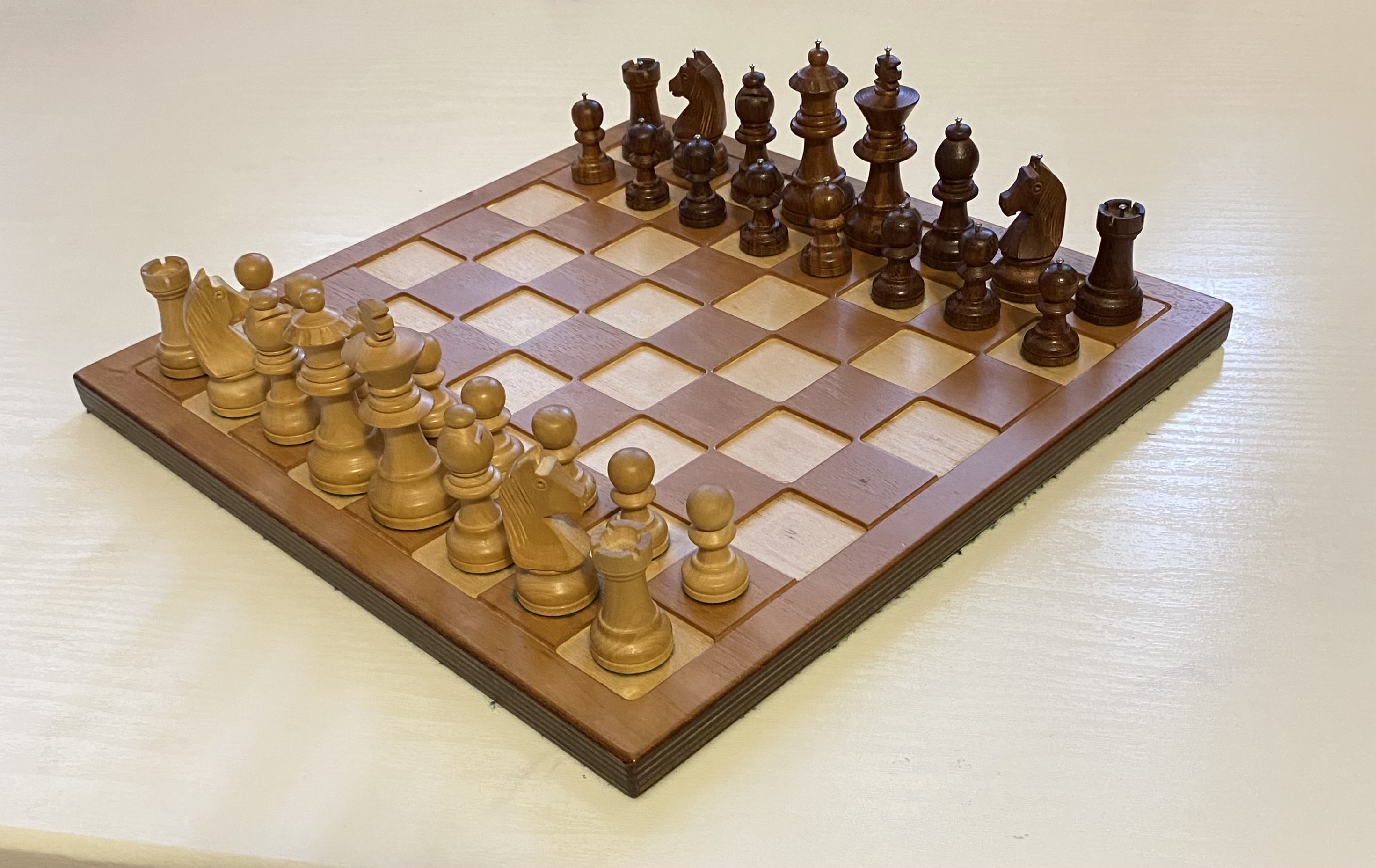 Scacchiera in legno per non vedenti, con pezzi degli scacchi posizionati nella disposizione iniziale per una partita. I pezzi chiari sono sulla sinistra e quelli scuri sulla destra. La scacchiera ha delle scanalature quadrate in cui si inseriscono i pezzi, per mantenerli stabili.